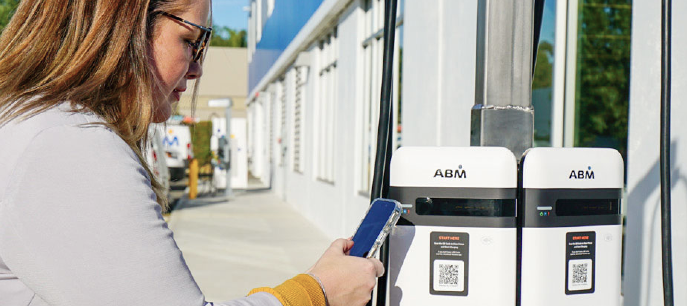 Woman wearing a white hard hat, safety glasses and an ABM shirt inspecting a newly installed EV charger.
